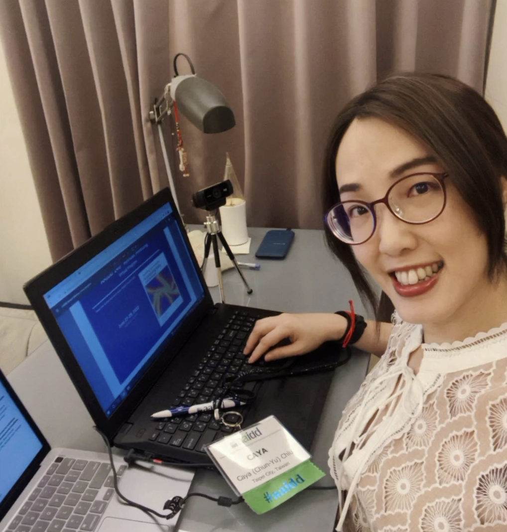 A smiling woman with dark brown hair and glasses takes a selfie at her desk, wearing a white cutout-patterned dress. Her workspace includes a black laptop displaying a blue presentation, a silver laptop to the left, a small tripod-mounted webcam, a smartphone, and a desk lamp. A conference badge rests on her keyboard reading 'aidd', 'CAYA', and 'Caya (Chun-Yu)', with a green '#aaidd' ribbon attached. Neutral curtains hang in the background.